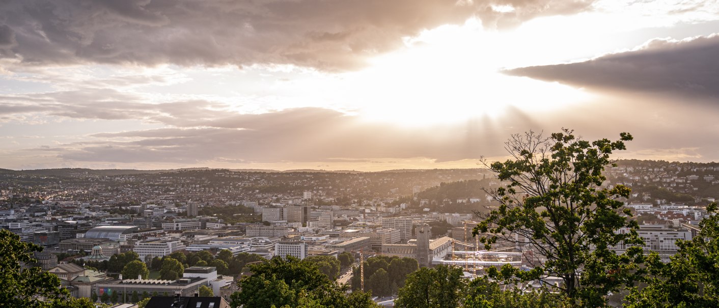 Sonnenuntergang über Stuttgart von der Uhlandshöhe aus. Die Stadt liegt unter einem bewölkten Himmel, durch den Sonnenstrahlen brechen., © Christine Garcia Urbina trickytine