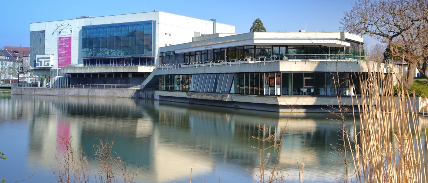 The Böblingen Congress Hall is reflected in the water, surrounded by bare trees and reeds, under a clear blue sky., © ccbs_gf