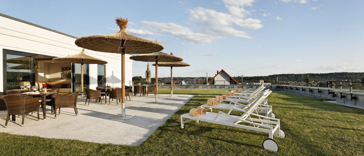 Terrasse mit Liegestühlen und Sonnenschirmen auf grünem Rasen, Blick auf eine Stadt im Hintergrund unter blauem Himmel., © Parkhotel Stuttgart Messe-Airport GmbH & Co KG Terrasse mit Liegestühlen und Sonnenschirmen auf grünem Rasen, Blick auf eine Stadt im Hintergrund unter blauem Himmel., © Parkhotel Stuttgart Messe-Airport GmbH & Co KG