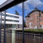 Moderne Glasfassade der Evangelischen Akademie Bad Boll spiegelt ein historisches Gebäude und den blauen Himmel wider. Im Vordergrund sind Stühle zu sehen., © Eabb_Giacinto_Carlucci
