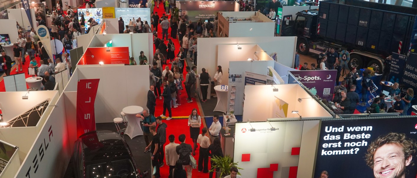 Menschen auf einer Messe mit zahlreichen Ständen, roten Teppichen und einem großen Werbebanner. Ein Tesla-Stand ist sichtbar., © Carl Benz Arena