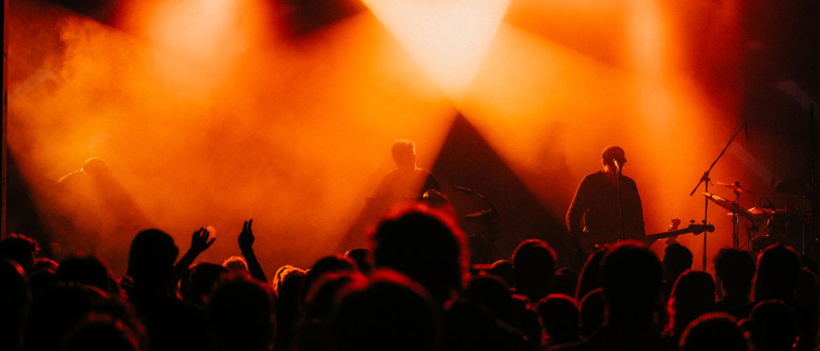 A band plays on a stage, illuminated by intense orange light, while the audience cheers in the foreground., &copy; Madlen Medvedovskyy