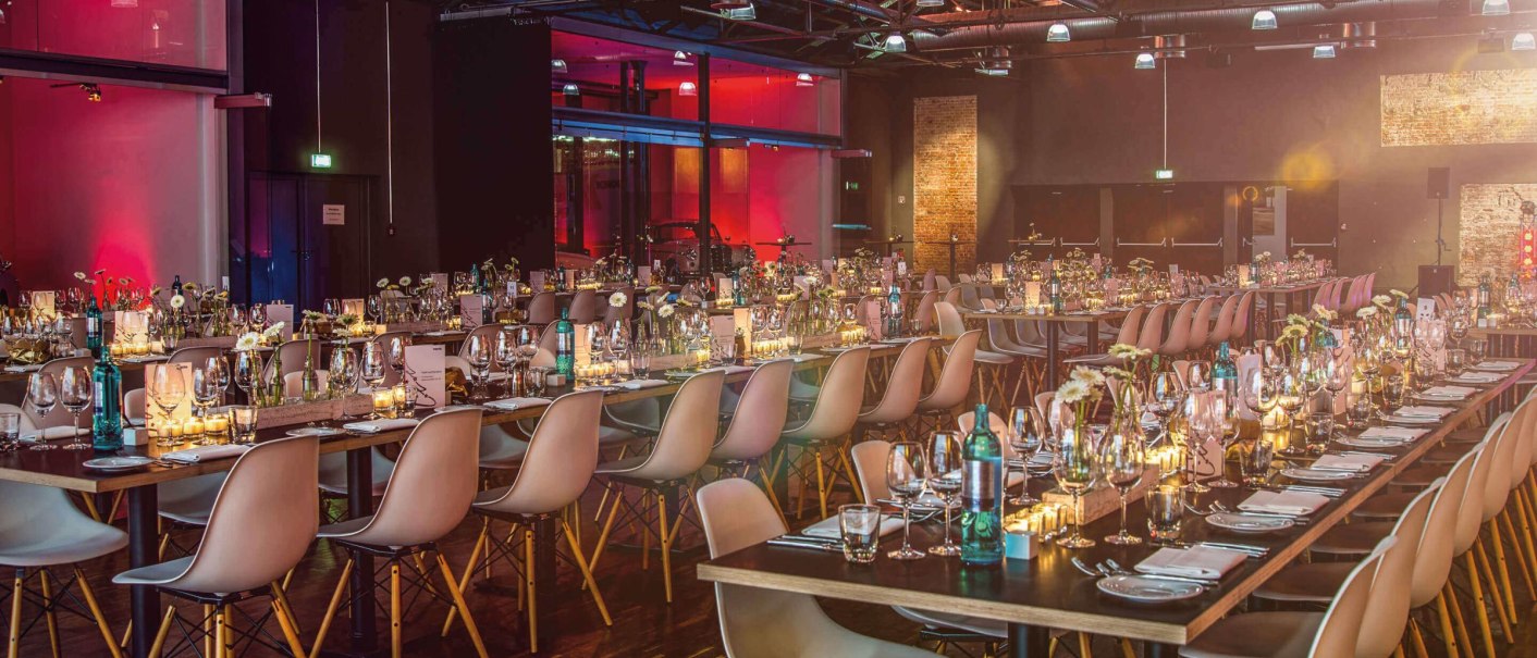 Elegante Halle mit langen, gedeckten Tischen und moderner Beleuchtung, bereit für ein Event. Die Atmosphäre ist warm und einladend., © rauschenbergercatering