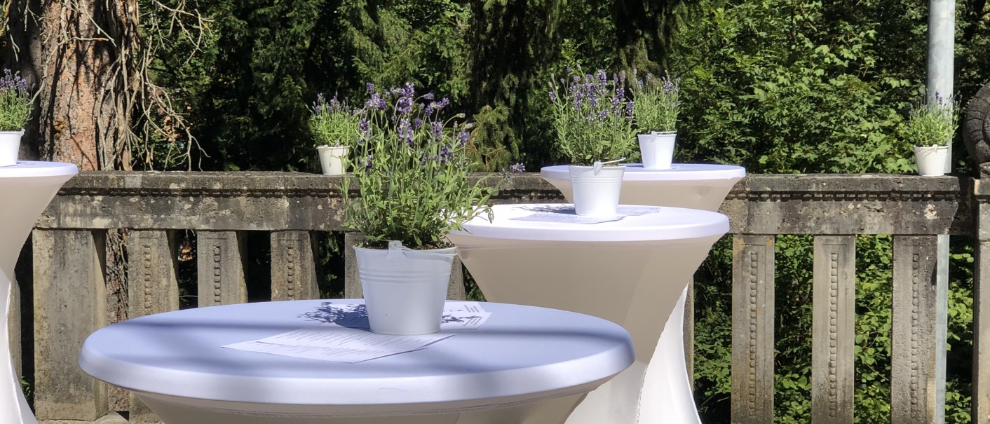 Stehtische mit weißen Tischdecken und Lavendelpflanzen auf einer Terrasse, umgeben von grünen Bäumen., © marmorsaalgmbh