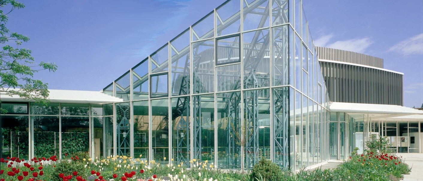 Modernes Glasgebäude der Stadthalle Sindelfingen, umgeben von bunten Blumenbeeten unter blauem Himmel., © ccbs