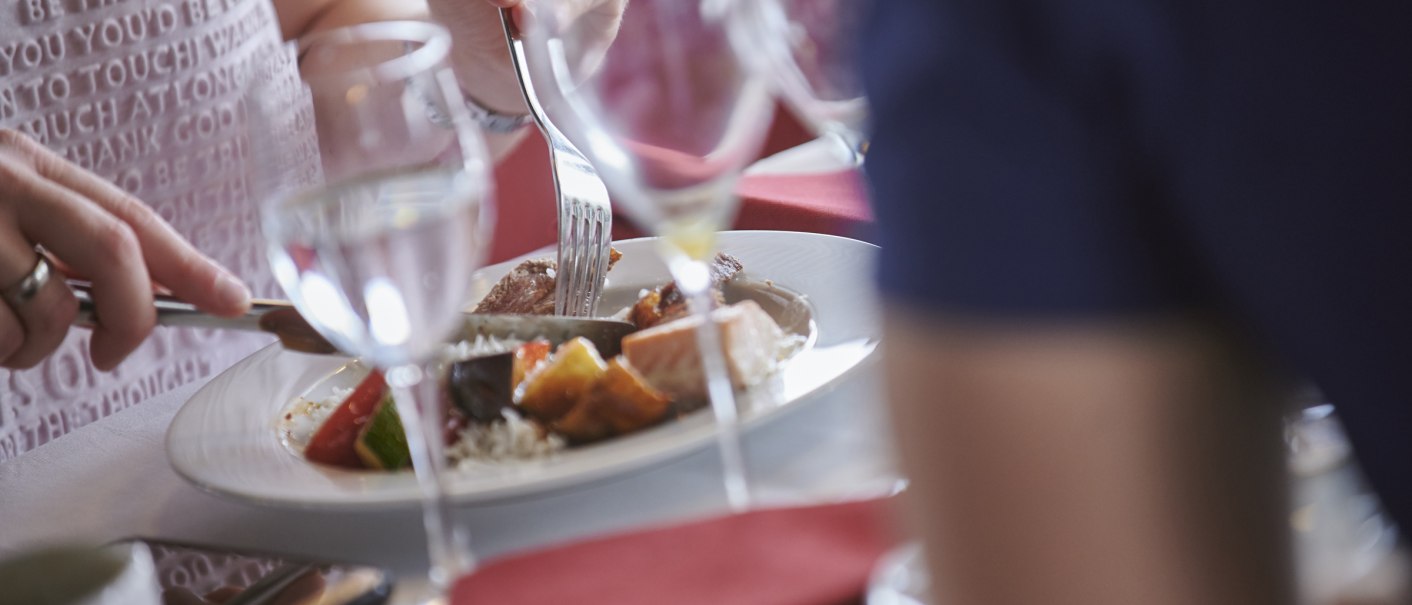 Personen essen an einem Tisch mit elegant gedecktem Geschirr. Ein Teller mit Fleisch und Gemüse ist zu sehen, umgeben von Gläsern und roten Servietten., © SWR Media Services GmbH / Martin Sigmund