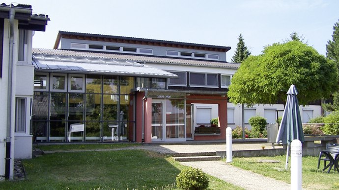 Modernes Geb&auml;ude mit Glasfassade, umgeben von Gr&uuml;nfl&auml;chen und einem Sonnenschirm. Klare Linien und gro&szlig;e Fenster pr&auml;gen die Architektur., &copy; L