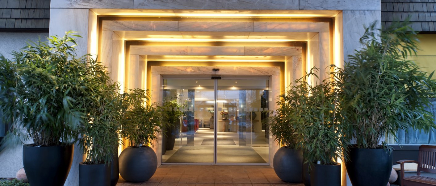 Modern hotel entrance with illuminated frame and revolving glass door, flanked by large plants in black pots., © Wyndham Stuttgart Airport Messe Modern hotel entrance with illuminated frame and revolving glass door, flanked by large plants in black pots., © Wyndham Stuttgart Airport Messe