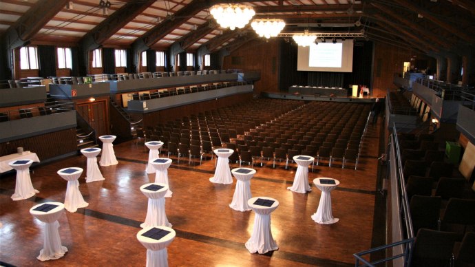 Interior view of the Stadthalle Herrenberg: empty rows of chairs, bar tables with white tablecloths, stage with projector, wooden floor and high ceiling., &copy; Stadt Herrenberg