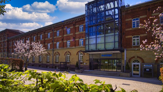 Das nestor Hotel Ludwigsburg zeigt eine Backsteinfassade mit moderner Glasfront. Blühende Bäume und blauer Himmel ergänzen die Szene., © nestor Hotel Ludwigsburg