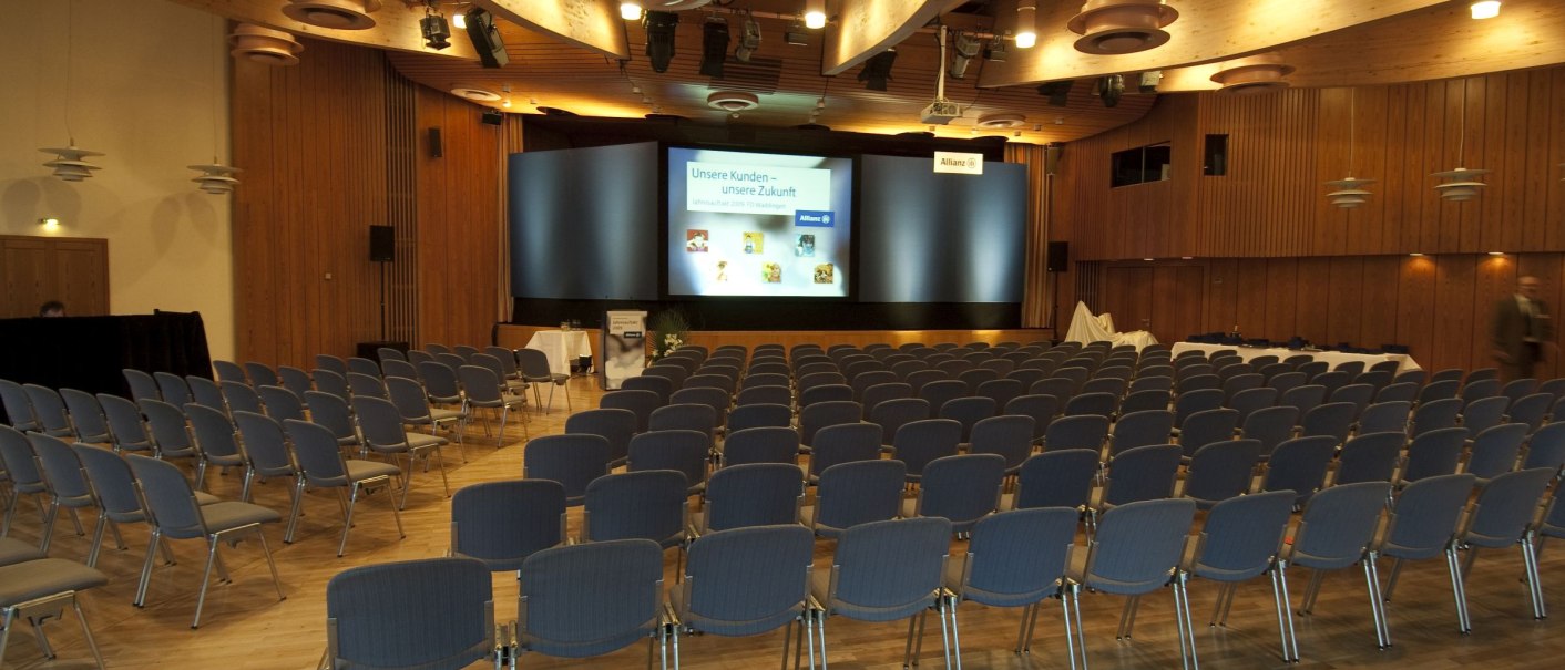 Empty conference room in the Waiblingen community center with rows of chairs and a stage. Wood paneling and modern lighting dominate the room., © Peter Oppenländer Empty conference room in the Waiblingen community center with rows of chairs and a stage. Wood paneling and modern lighting dominate the room., © Peter Oppenländer