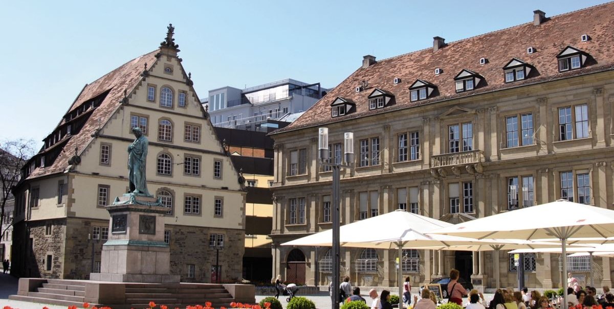 Schillerplatz in Stuttgart mit Fruchtkasten, Schillerdenkmal und Menschen in einem Café. Bunte Blumen im Vordergrund, blauer Himmel im Hintergrund., © Stuttgart-Marketing GmbH