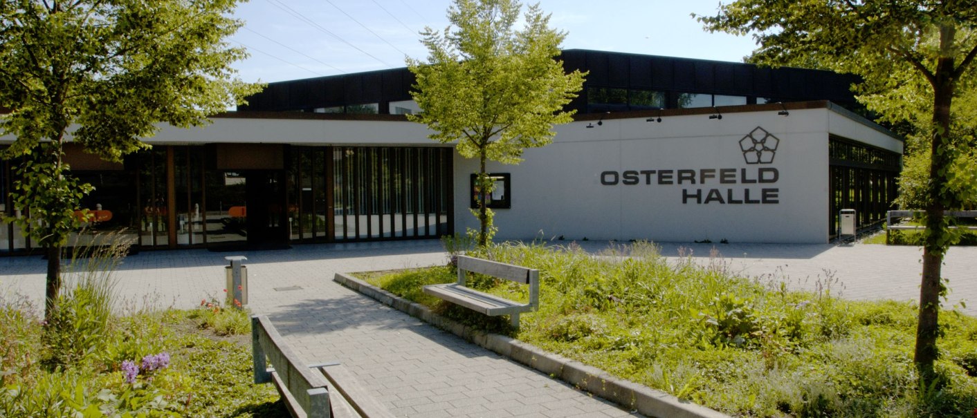 Die Osterfeldhalle von außen, umgeben von Bäumen und Pflanzen. Ein gepflasterter Weg führt zu Bänken vor dem Gebäude., © eslive Die Osterfeldhalle von außen, umgeben von Bäumen und Pflanzen. Ein gepflasterter Weg führt zu Bänken vor dem Gebäude., © eslive