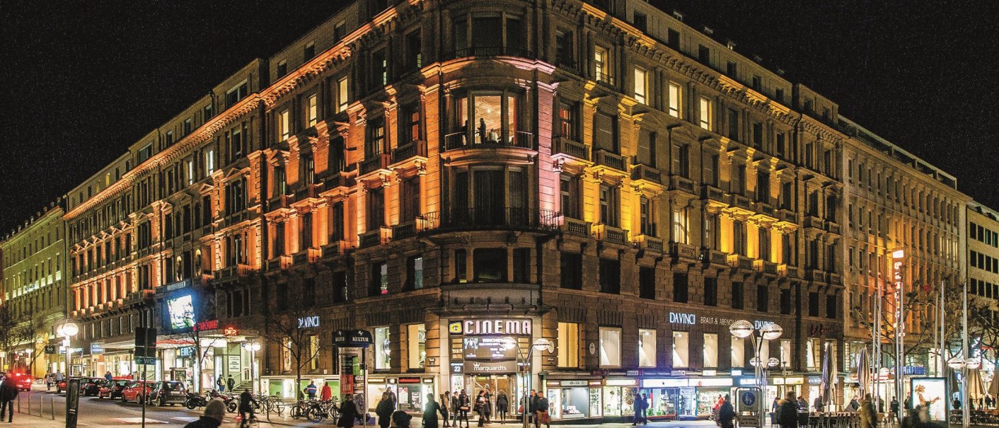 Ein beleuchtetes Gebäude bei Nacht mit einem Kino und Geschäften im Erdgeschoss. Menschen gehen auf der Straße vorbei., © Schauspielbühnen Stuttgart