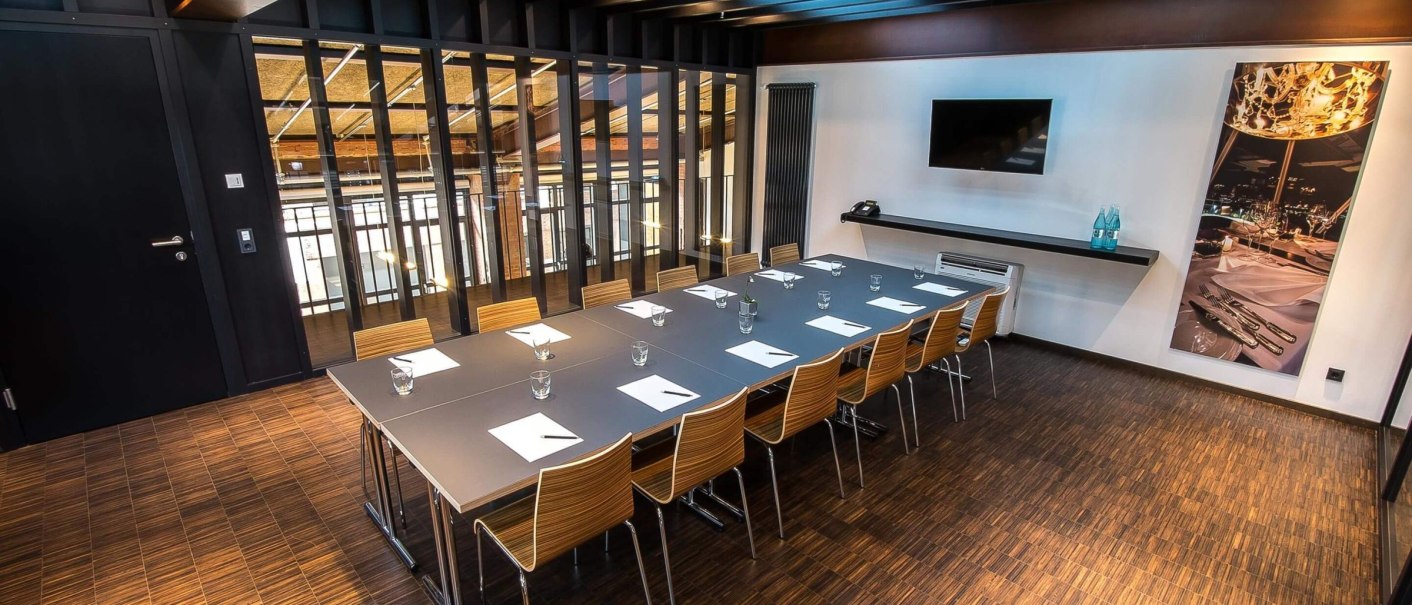 Moderner Meetingraum mit großem Tisch, Stühlen, TV an der Wand und dekorativem Bild. Der Raum hat eine Holzoptik und ist stilvoll eingerichtet., © rauschenbergercatering