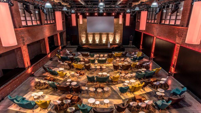 Modern event room with colorful armchairs and tables, stage and screen. Brick walls and warm lighting create an inviting atmosphere., © rauschenbergercatering