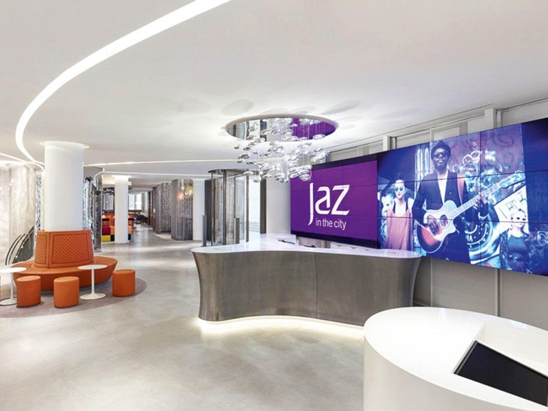 Modern hotel reception with curved counter, large screen with 'Jaz in the City' logo and stylish lighting., &copy; Jaz Hotel Stuttgart