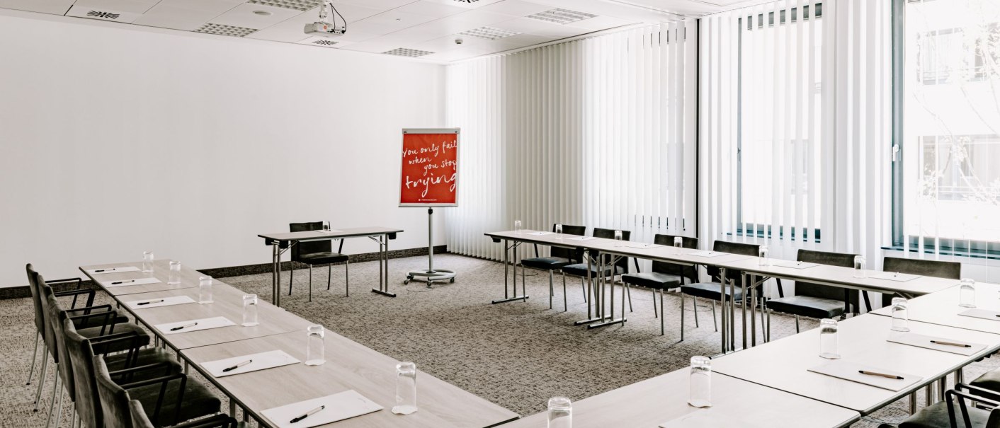 Bright meeting room with U-shaped table arrangement, chairs, notepads and pens. A flipchart with a motivational quote stands at the front., &copy; Vienna House Easy Mo. Stuttgart