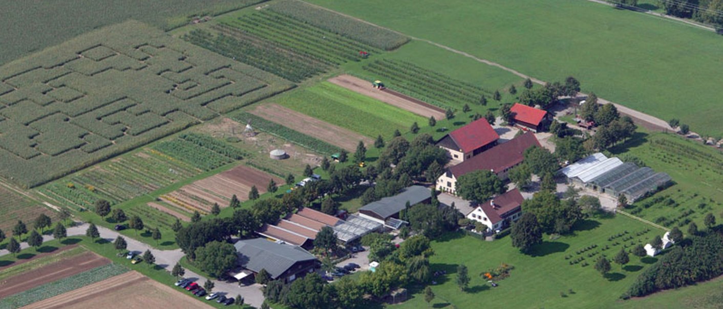 Luftaufnahme eines Bauernhofs mit mehreren Gebäuden, Gewächshäusern und Feldern. Ein Labyrinth ist in einem Feld zu erkennen., © Sonnenhof GmbH & Co KG Luftaufnahme eines Bauernhofs mit mehreren Gebäuden, Gewächshäusern und Feldern. Ein Labyrinth ist in einem Feld zu erkennen., © Sonnenhof GmbH & Co KG