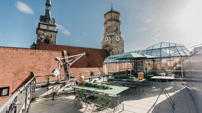 Dachterrasse mit gr&uuml;nen Tischen und St&uuml;hlen, umgeben von Glasstrukturen. Im Hintergrund sind zwei Kircht&uuml;rme zu sehen, unter blauem Himmel., &copy; OutOfOffice GmbH