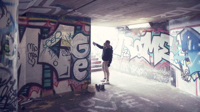 Eine Person sprüht Graffiti in einem Tunnel. Die Wände sind mit bunten Graffitis bedeckt. Auf dem Boden stehen Farbdosen., © hoponopo GmbH Eine Person sprüht Graffiti in einem Tunnel. Die Wände sind mit bunten Graffitis bedeckt. Auf dem Boden stehen Farbdosen., © hoponopo GmbH