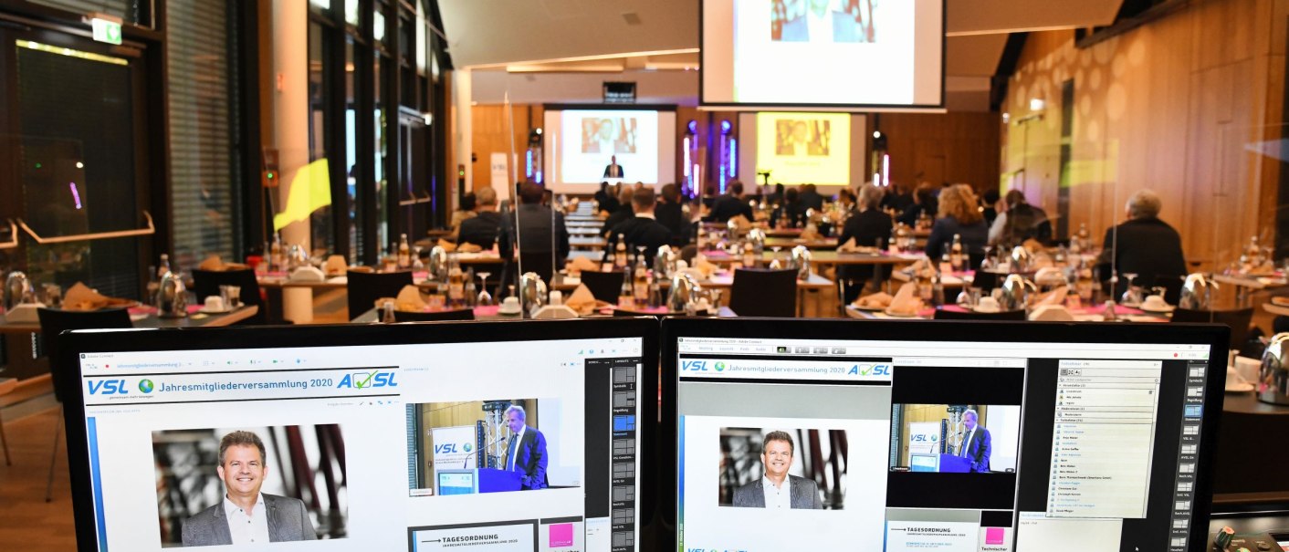 Hybride Tagung in der Filderhalle: Zwei Bildschirme im Vordergrund zeigen Präsentationen, während im Hintergrund Teilnehmer an Tischen sitzen und einem Vortrag folgen., © Guenter E. Bergmann - Photography