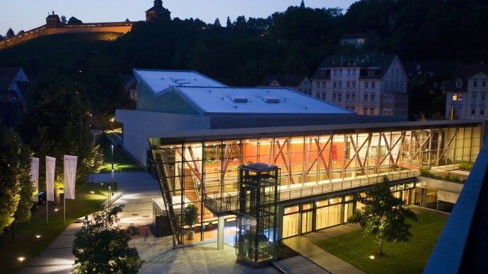 Das Neckar Forum bei Nacht, ein modernes Glasgebäude, ist hell erleuchtet. Im Hintergrund ist eine beleuchtete Burg auf einem Hügel zu sehen., © eslive_Thomas Wagner