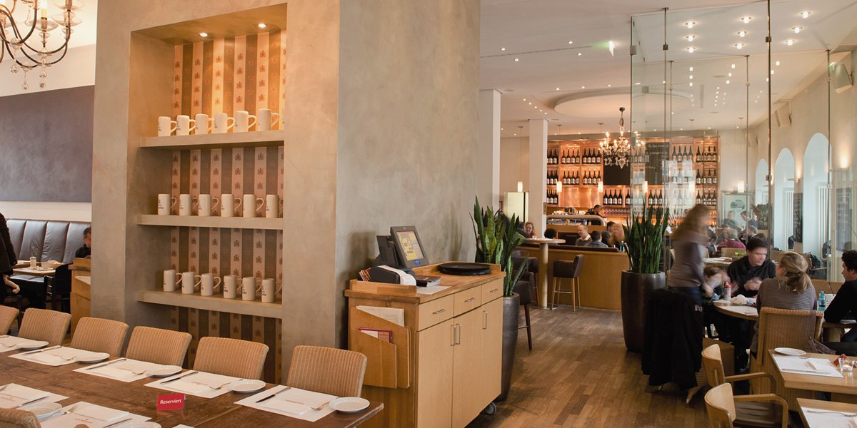 Stylish restaurant with wooden furniture, shelves full of cups and a cozy atmosphere. Guests sit at tables, with a bar in the background., &copy; Aspectus Photographie Stuttgart