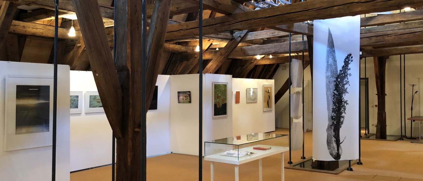 Kunstausstellung in einem Raum mit Holzbalken, weißen Wänden und verschiedenen Kunstwerken. Ein Glastisch steht in der Mitte., © kkmaisch_gf