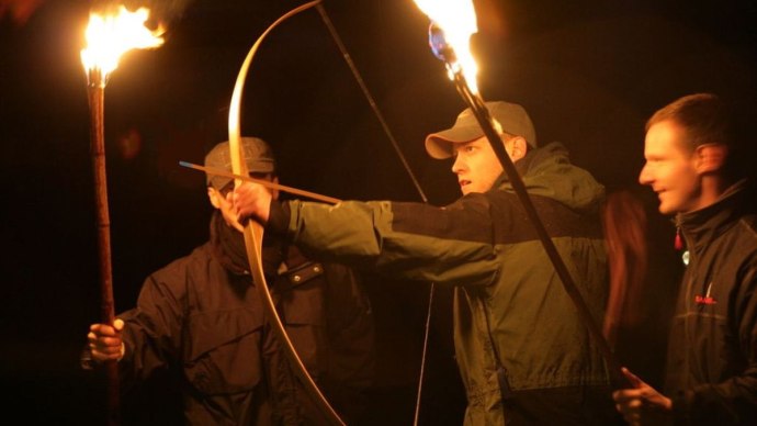 Drei Männer schießen nachts mit Bögen, beleuchtet von Fackeln. Einer spannt den Bogen, während die anderen zuschauen. Dunkle Umgebung., © Teamgeist Süd GmbH Drei Männer schießen nachts mit Bögen, beleuchtet von Fackeln. Einer spannt den Bogen, während die anderen zuschauen. Dunkle Umgebung., © Teamgeist Süd GmbH