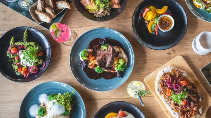 Ein Tisch mit verschiedenen Gerichten: Fleisch mit Gemüse, Salate, Burger mit Pommes, Brot und Desserts. Getränke sind ebenfalls zu sehen., © SatzDrei Ein Tisch mit verschiedenen Gerichten: Fleisch mit Gemüse, Salate, Burger mit Pommes, Brot und Desserts. Getränke sind ebenfalls zu sehen., © SatzDrei