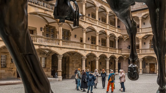 Innenhof eines alten Schlosses mit Arkaden und Säulen. Eine Gruppe von Menschen steht zusammen, im Vordergrund sind die Beine einer Pferdestatue sichtbar., © Stuttgart Marketing GmbH - Sarah Schmid Innenhof eines alten Schlosses mit Arkaden und Säulen. Eine Gruppe von Menschen steht zusammen, im Vordergrund sind die Beine einer Pferdestatue sichtbar., © Stuttgart Marketing GmbH - Sarah Schmid