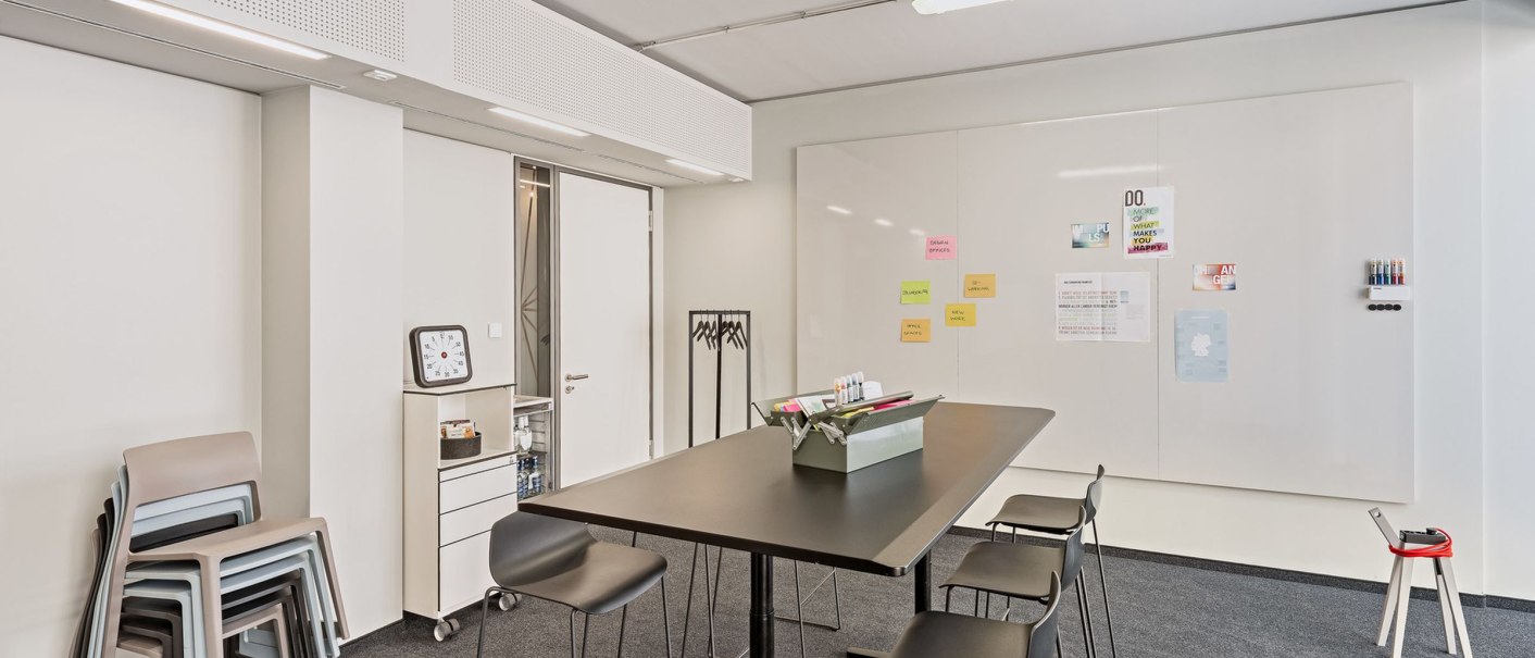 Moderner Besprechungsraum mit hohem Tisch, schwarzen Stühlen und einem Whiteboard mit Notizen. Stapelbare Stühle und eine Uhr sind ebenfalls sichtbar., © Design Offices Moderner Besprechungsraum mit hohem Tisch, schwarzen Stühlen und einem Whiteboard mit Notizen. Stapelbare Stühle und eine Uhr sind ebenfalls sichtbar., © Design Offices