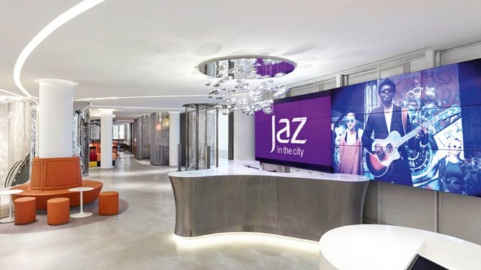Moderne Hotelrezeption mit geschwungenem Tresen, großem Bildschirm mit 'Jaz in the City' Logo und stilvoller Beleuchtung., © Jaz Hotel Stuttgart