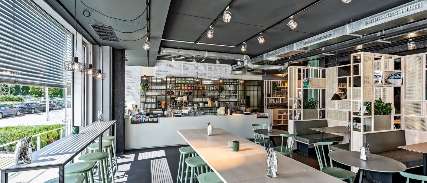 Modernes Café mit langen Tischen, grünen Stühlen und großen Fenstern. Im Hintergrund eine Theke mit Regalen und Speisekarte., © Design Offices GmbH