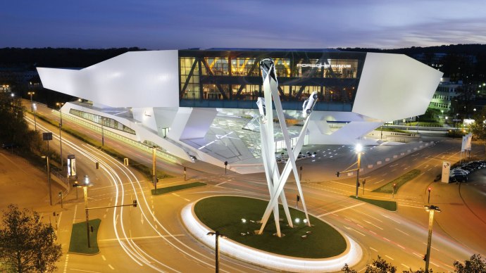 The Porsche Museum in Stuttgart at night, with modern architecture and illuminated streets. A sculpture of Porsche vehicles in the foreground., © Porsche AG