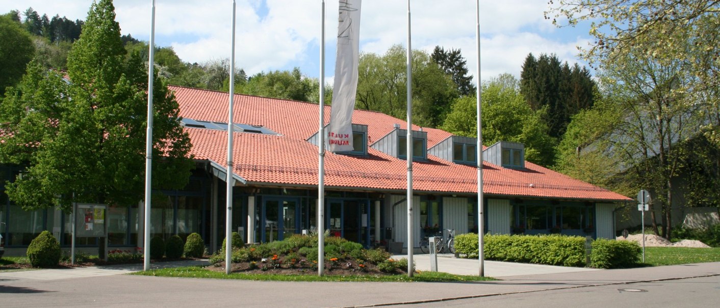 The Murrhardt town hall with its red tiled roof, surrounded by trees and flagpoles, on a sunny day., © Stadt Murrhardt