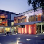 Die Stadthalle K3N bei Dämmerung, moderne Glasarchitektur mit beleuchtetem Vorplatz und Fahnenmasten., © k3nmaisch