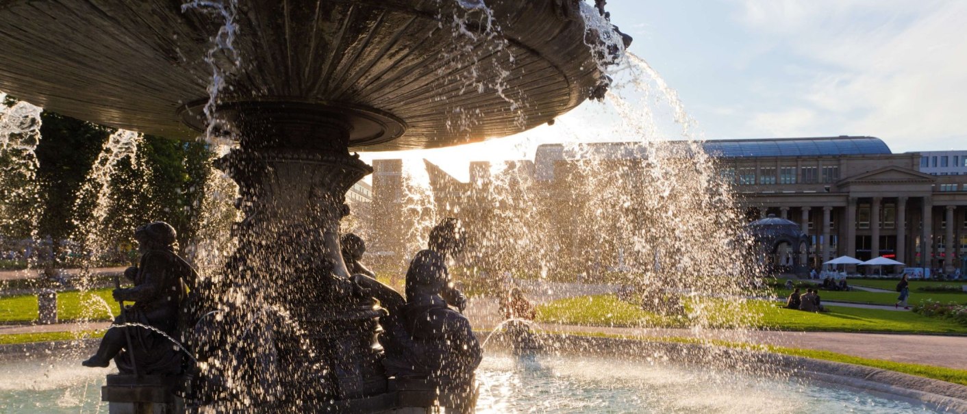 Ein Brunnen am Schlossplatz mit sprudelndem Wasser im Vordergrund. Im Hintergrund sind historische Geb&auml;ude und Menschen auf einer Wiese zu sehen., &copy; Werner Dieterich