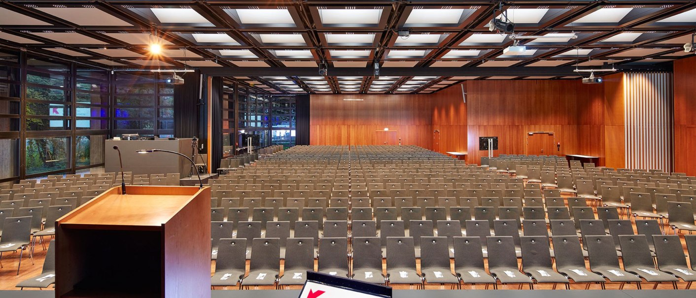 Leerer Festsaal mit Stuhlreihen, Rednerpult und Bildschirm mit 'Herzlich Willkommen'. Holzwände und moderne Deckenbeleuchtung., © Steffen Scheyhing Fotodesignwww.steffenscheyhing.de