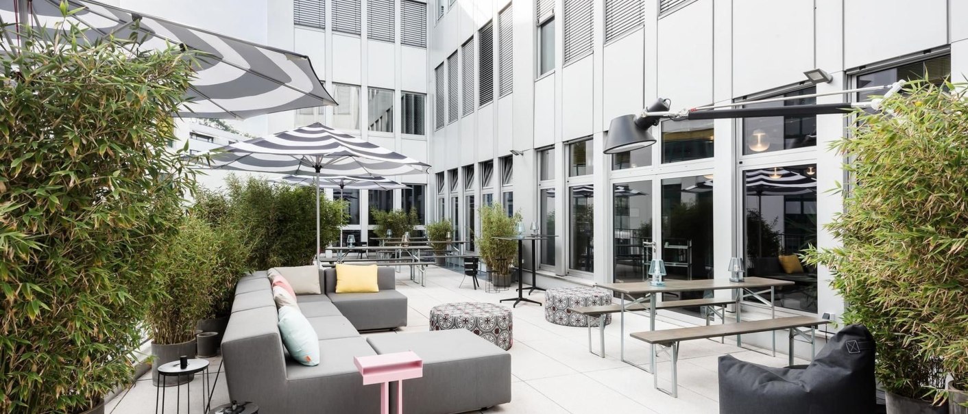 Moderne Terrasse mit grauen Sofas, bunten Kissen, Sonnenschirmen und Pflanzen. Helle Fassade im Hintergrund. Design Offices Stuttgart Tower Terrasse., © Design Offices GmbH