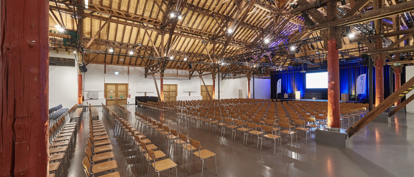 Große Veranstaltungshalle mit Holzdecke, vielen Stuhlreihen und Bühne. Die Beleuchtung ist warm, und die Architektur zeigt sichtbare Holzbalken., © PeterHartung Große Veranstaltungshalle mit Holzdecke, vielen Stuhlreihen und Bühne. Die Beleuchtung ist warm, und die Architektur zeigt sichtbare Holzbalken., © PeterHartung